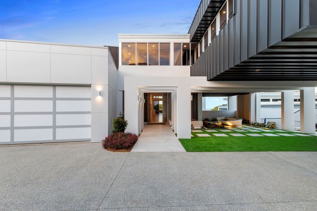 Exterior view of a contemporary multi-level residence with a white rendered facade, large integrated garage, projecting upper-level balcony or overhang, and dark vertical cladding. A covered entry walkway leads through the centre, with a landscaped courtyard and geometric lawn visible beneath the cantilevered upper floor.