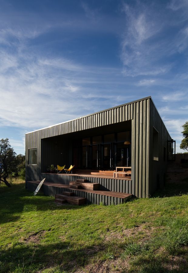 Exterior photograph of a compact contemporary single-storey house set in a grassy landscape. The building has a strongly articulated dark vertical-ribbed cladding, a steep asymmetric pitched roof, extensive recessed glazing, and a raised timber deck with broad timber steps. Outdoor chairs and a small bench sit on the covered veranda.
