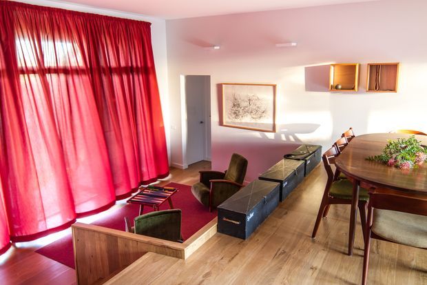A bright, mid-century-inspired open-plan living and dining area. The room features dramatic floor-to-ceiling red curtains enclosing a slightly sunken lounge zone, warm timber flooring and built-in timber elements, a dark low media console, vintage-style upholstered lounge chairs, and a large rounded timber dining table with chairs. White walls, minimalist wall-mounted lighting, framed artwork, and compact timber display niches complete the interior.