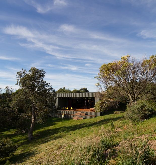 A low, single-storey modern house set into a gently sloping, densely planted landscape. The dark rectangular facade has a broad corrugated-looking roof and deep covered outdoor living area, opening onto a timber deck with steps and bright yellow seating. Mature trees, grasses and shrubs surround the dwelling beneath a wide blue sky.