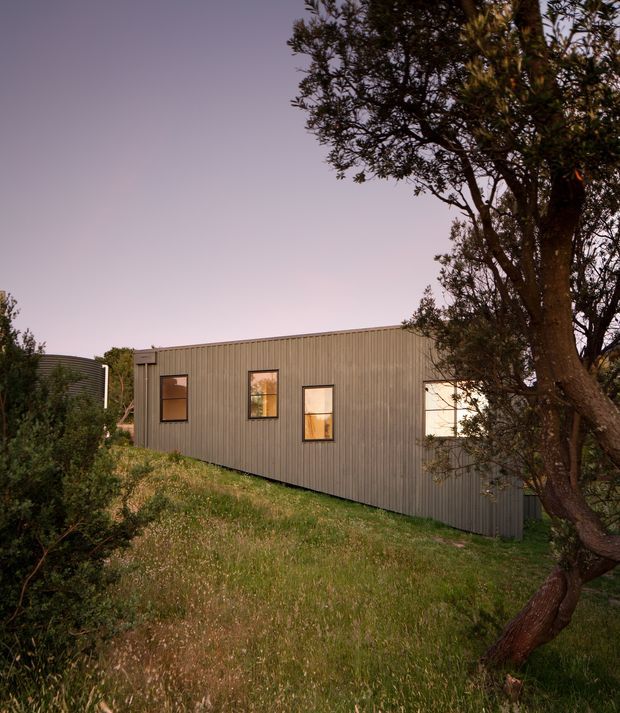 A compact single-storey building viewed from a grassy landscaped slope at dusk. The long rectangular facade is clad in vertically ribbed grey metal with several warm-lit rectangular windows, while mature trees and shrubs frame the structure. The simple low-pitched or flat roofline and restrained material palette give the exterior a contemporary appearance.