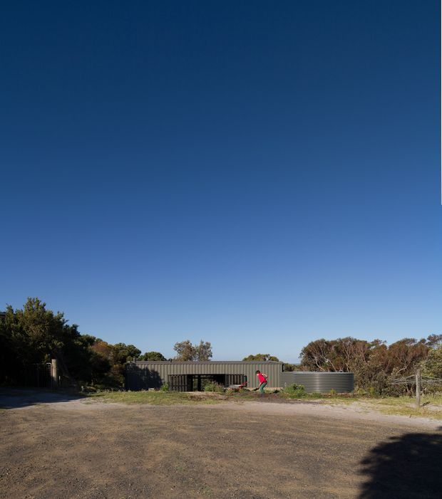 A low, elongated contemporary building or outbuilding set behind a broad gravel forecourt, with dark corrugated metal cladding, a shallow pitched roof, and a large glazed or screened opening. Native-looking vegetation and trees surround the structure under a clear blue sky. A person stands near the facade, providing scale.