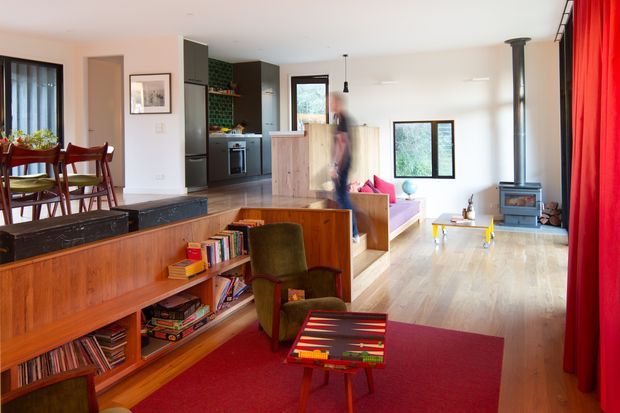 Open-plan mid-century-inspired living area with a dining zone at left, a sunken or stepped living room in the centre, and a compact kitchen toward the rear. Warm timber built-in cabinetry and shelving define the room, with a red area rug and full-height red curtains adding bold colour. A freestanding wood-burning stove, pale timber flooring, exposed dark kitchen cabinetry, and large windows create a bright, lived-in interior.