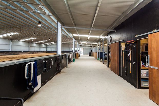 Wide interior view of a large equestrian or agricultural facility, with a concrete circulation aisle, dark timber and black storage partitions, open stalls or work bays on the left, and exposed metal roof framing with translucent roof panels and industrial lighting.