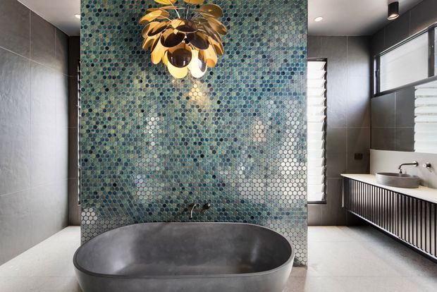Contemporary bathroom featuring a freestanding dark grey bathtub in front of a full-height iridescent blue-green mosaic tile feature wall. Large grey wall tiles, pale flooring, a floating vanity with a white vessel basin and dark vertical slatted cabinetry, louvred windows, and a sculptural gold pendant light create a refined, high-contrast scheme.