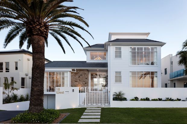 Front exterior view of a refined multi-storey coastal-style residence with white painted weatherboard walls, dark tiled pitched roofs, large white-framed windows, and a gated entry. A palm-lined lawn, concrete stepping-stone path, low white boundary wall, and landscaped planting frame the facade.