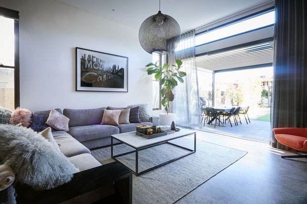 Bright contemporary living room with a large grey sectional sofa, textured cushions, black-framed glass coffee table, woven area rug, pendant light, indoor plant, and a vivid orange lounge chair. Full-height glazing and sliding doors open toward an outdoor terrace, with a dining area visible beyond.
