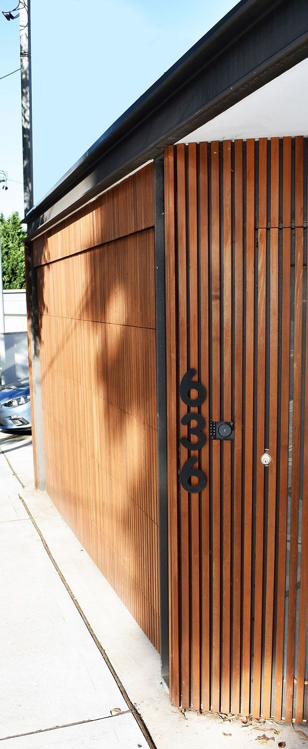 Close-up exterior view of a modern timber-clad entry gate and adjoining wall. Warm vertical wooden slats are framed with dark metal, with a black geometric house number, keypad, and lock hardware. A narrow concrete walkway runs alongside the facade, and a simple dark roof edge is visible above.