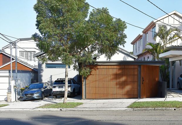 Street-facing view of a contemporary multi-storey residential property with a broad timber garage door and matching timber pedestrian gate beneath a dark horizontal canopy. White rendered walls, projecting balconies, vertical cladding, large glazing, palm planting and a paved driveway are visible, with neighbouring homes and parked cars in the surrounding suburban streetscape.