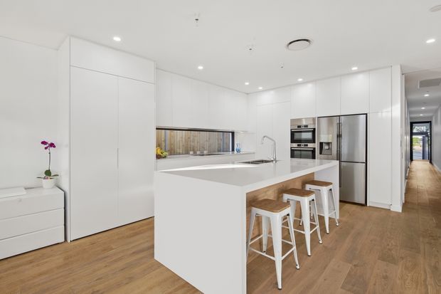 Bright, contemporary white kitchen with full-height handleless cabinetry, a large white island with integrated sink and seating for four, stainless-steel appliances, and warm timber-look flooring. A narrow horizontal window provides a view to timber screening, while a hallway extends beyond the kitchen.