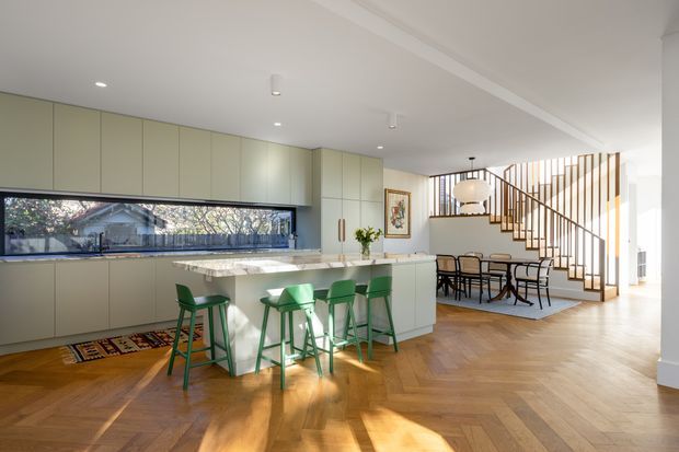 A bright open-plan kitchen and dining area with pale green full-height cabinetry, a light stone island, and four green bar stools. The room features warm herringbone timber flooring, a long horizontal window above the benchtop, and a dining table positioned beneath a sculptural pendant light. A timber staircase with vertical balustrade screening rises behind the dining area, contributing a refined contemporary architectural character.