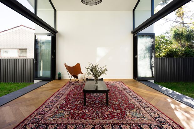 A bright, minimalist enclosed sunroom or indoor-outdoor living space with full-height black-framed glazing and open glass doors on both sides. The room has a warm herringbone timber floor, a large ornate Persian-style rug, a low dark coffee table with a vase of branches, and a sculptural brown leather lounge chair. Black corrugated fencing and neighbouring residential architecture are visible outside.