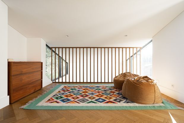 Bright minimalist upper-level lounge or sitting area with a white ceiling and walls, warm herringbone timber flooring, a colourful geometric area rug, two brown leather beanbag-style seats, and a low wooden chest of drawers. Full-height glazing and a vertical timber slat balustrade create an airy connection to the adjacent space.