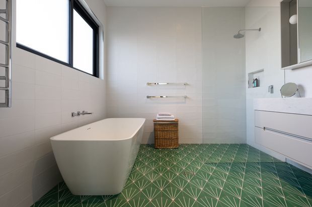 Bright contemporary bathroom featuring a freestanding white bathtub, green geometric patterned floor tiles, pale wall tiles, a wall-mounted white vanity with integrated basin, frameless shower glazing, and black-framed windows. The room has clean lines, abundant natural light, and restrained modern fixtures.