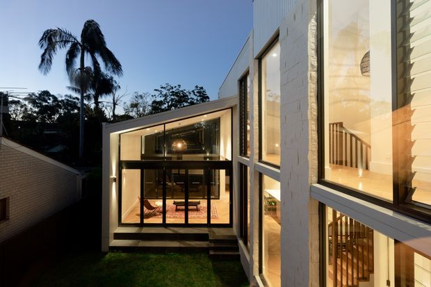 Twilight exterior view of a contemporary multi-level residence, focused on a glazed central living area and a tall side facade with expansive windows. Warm interior lighting reveals a stairway, upper-level workspace or living area, and double-height glazing. White masonry walls, dark window frames, clean geometric volumes, and a small lawn create a refined modern composition, with palm trees silhouetted in the background.
