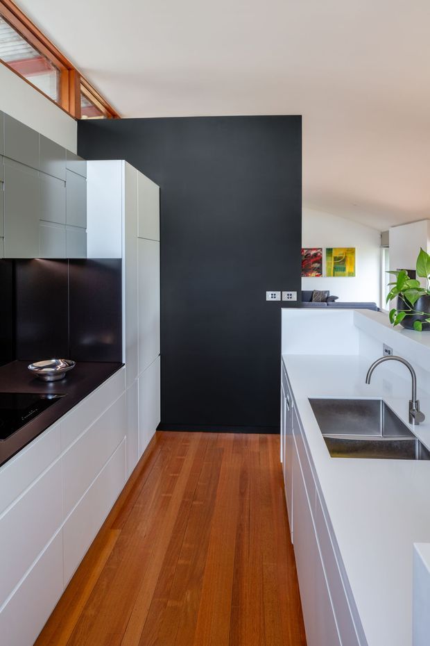 A narrow, contemporary galley kitchen with white handleless cabinetry, a long white island or workbench, stainless-steel double sink and tap, dark stone-look benchtops and a dramatic charcoal feature wall. Warm timber flooring and exposed timber-framed clerestory glazing add contrast, while a glimpse of the adjoining living area is visible beyond the kitchen.