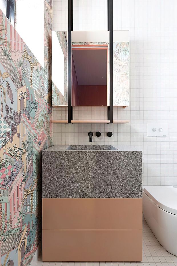 Compact contemporary bathroom featuring a terrazzo vanity with integrated basin, pale peach cabinetry, white small-format tiled walls and floor, a freestanding toilet, and a bold illustrated patterned feature wall. A black-framed mirrored cabinet and dark tapware provide contrast, while a narrow skylit vertical opening brings in natural light.