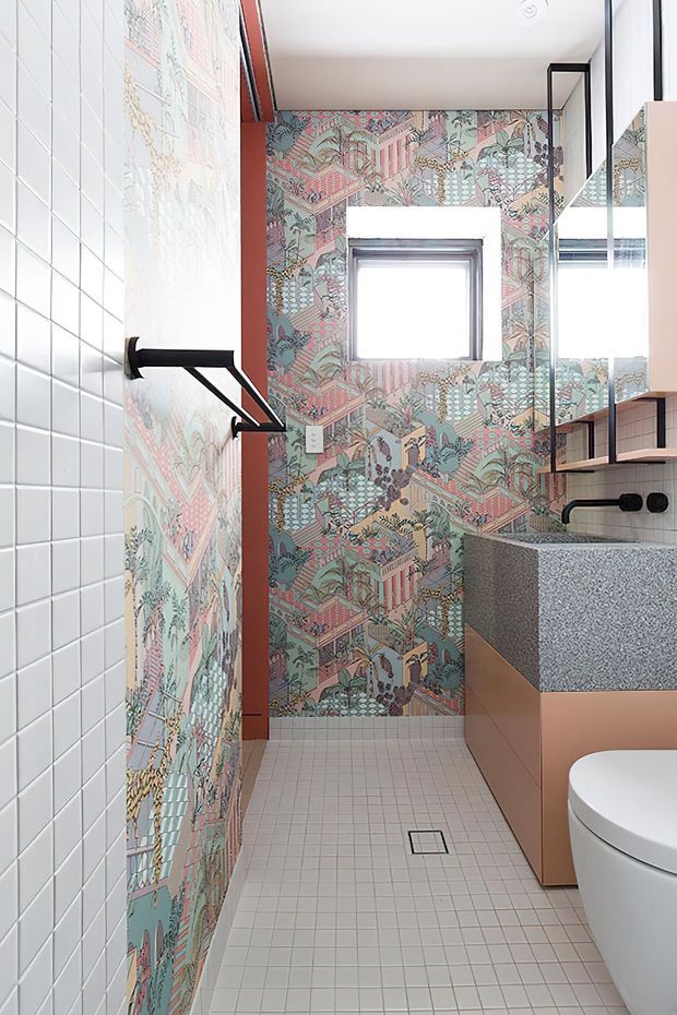 A compact contemporary bathroom with a walk-in shower, wall-mounted black towel rail, patterned feature wallpaper, white square wall tiles, pale pink grid floor tiles, and a floating timber-toned vanity with a speckled grey benchtop. A mirrored cabinet and toilet are visible on the right, with bright natural light through the window.