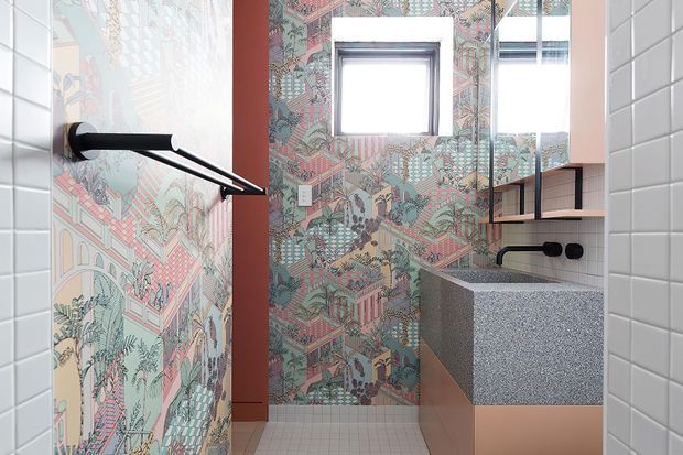 A compact contemporary bathroom featuring white tiled side walls, a colourful illustrated feature-tile wall, a black wall-mounted towel rail, a small square window, a mirrored cabinet, open shelving, and a terrazzo-like grey vanity benchtop with warm blush cabinetry.