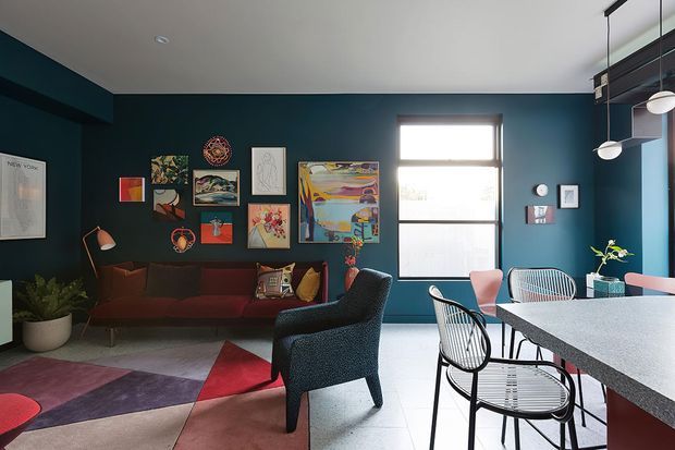 A colourful contemporary open-plan living and dining space with deep teal walls, a dark red sofa, patterned area rug, and a gallery wall of eclectic artwork. The dining area features a large stone-look table, wire-frame chairs, pendant lighting, and a bright window, creating a bold mid-century-inspired interior.