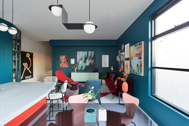 A vibrant contemporary open-plan living and dining area with a large island or dining table in the foreground, colourful upholstered chairs, a deep teal feature wall, extensive artwork, and sculptural pendant lighting. The room includes a lounge seating area, built-in cabinetry, dark-framed glazing, and a bold mix of red, pink, teal, and neutral finishes.