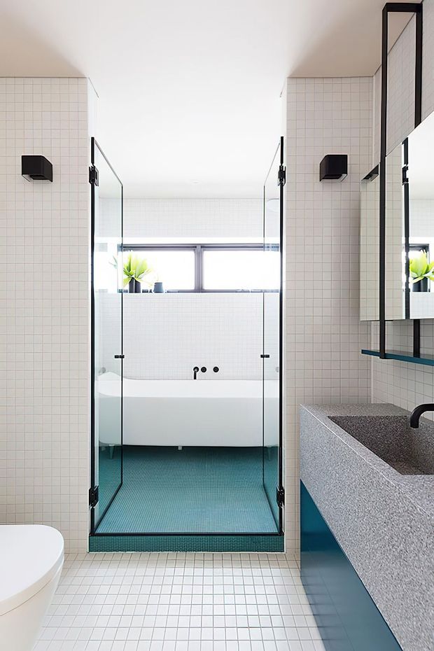Bright contemporary bathroom featuring a glass-enclosed walk-in shower, freestanding white bathtub, wall-hung toilet, large mirrored cabinet, and a blue integrated vanity with a speckled stone benchtop. Small white grid tiles line the walls and floor, with a turquoise shower floor and black fixtures.