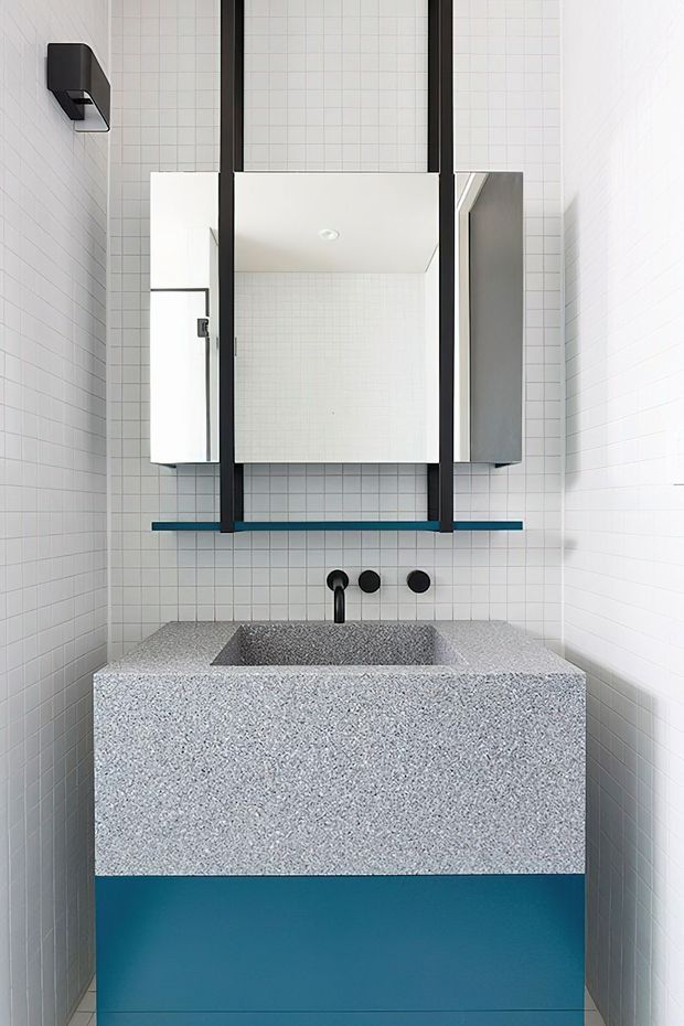A compact contemporary bathroom featuring white small-format tiled walls, a terrazzo-like grey vanity with integrated basin, teal-blue lower cabinetry or panel, black wall-mounted tapware, and a large black-framed mirrored cabinet above the basin.
