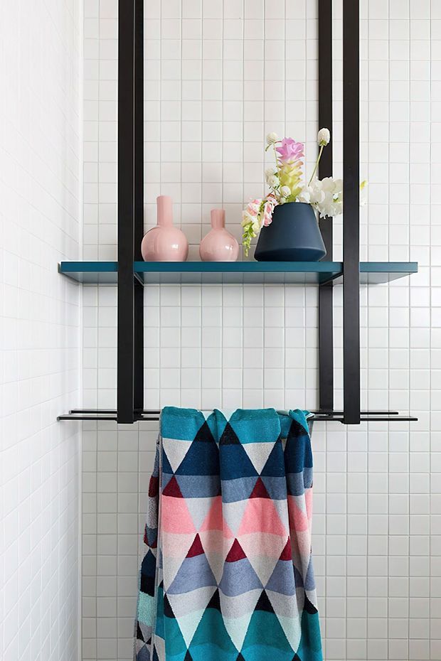 A bright, compact bathroom corner with white square tiled walls, a black metal-framed open shelving unit, and a teal shelf displaying pastel ceramic vases and flowers. A colourful geometric towel hangs from a lower black rail, adding a playful accent to the clean tiled setting.