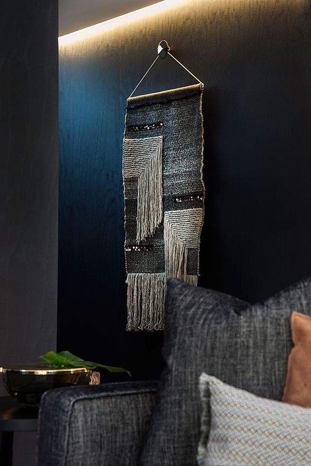 A moody interior detail showing a dark textured wall with concealed linear lighting, a handcrafted woven textile wall hanging, and the corner of a charcoal upholstered sofa with layered cushions. A small metallic side table with a green plant sits in the foreground.