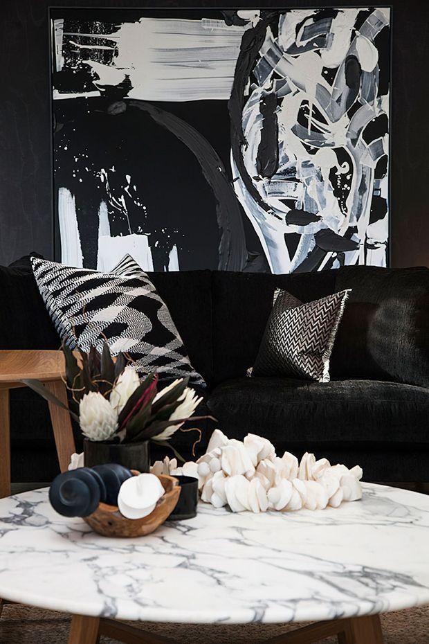A sophisticated living room vignette featuring a black velvet sofa with monochrome patterned cushions, a large black-and-white abstract portrait artwork, and a round white marble coffee table. Decorative objects, sculptural white pieces, and a floral arrangement create a dramatic monochromatic scheme with warm timber accents.