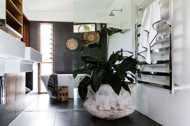 Contemporary bathroom with a freestanding white bathtub, wall-mounted fixtures, large-format dark floor and wall tiles, a heated towel rail, built-in timber shelving, and a prominent sculptural stone planter filled with lush tropical foliage. Natural light enters through a narrow timber-framed window, creating a refined indoor-outdoor feel.