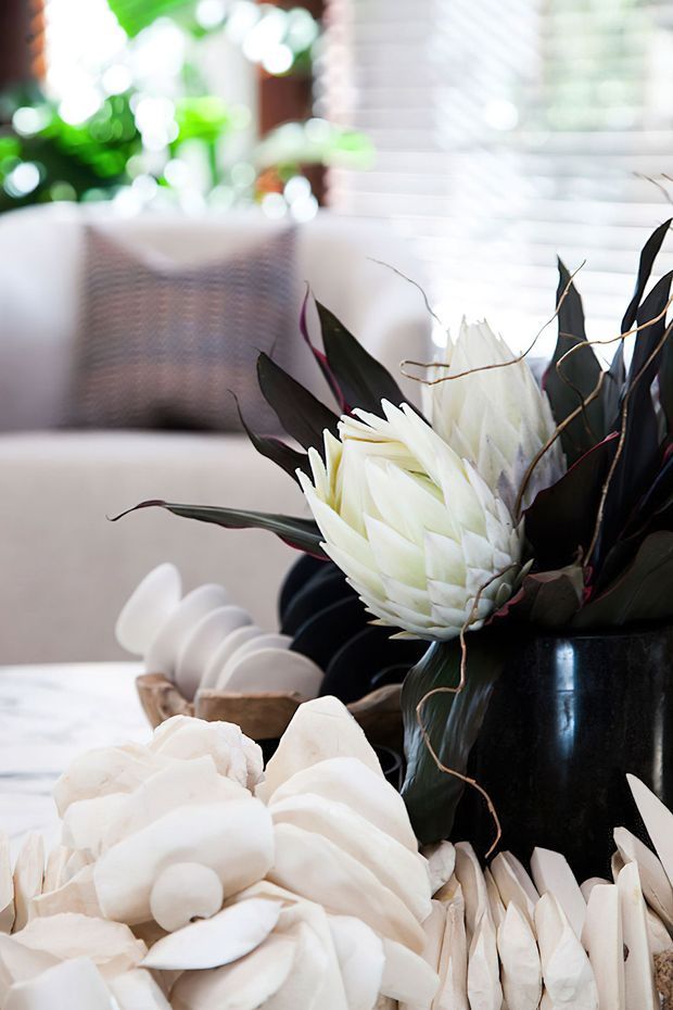 Close-up interior styling vignette featuring a large arrangement of white protea-like flowers and dark foliage in a glossy black vessel, with pale decorative objects on a light table in the foreground. A soft neutral sofa, woven cushion, bright window blinds, and blurred green plants create a bright, refined living-room setting.