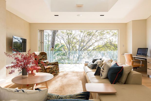 Bright, warm contemporary living room with a large floor-to-ceiling glazed opening overlooking leafy trees. The room features a cream sectional sofa, patterned cushions, low rounded timber coffee tables, a textured rug, sculptural lounge chairs, a wall-mounted television, and a compact desk workspace. Light neutral walls and abundant natural light create an airy, relaxed atmosphere.