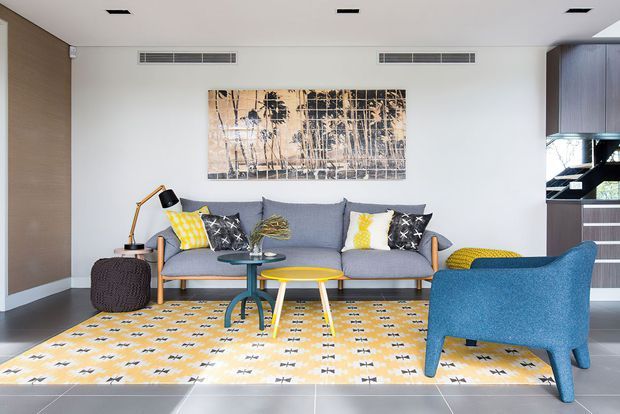 Bright contemporary living room with a grey sofa, blue armchair, yellow patterned rug, colourful cushions, nested coffee tables, and abstract wall art. The room has a clean white ceiling with recessed vents and lighting, dark tiled flooring, timber-toned wall panelling, and a staircase visible at the rear.