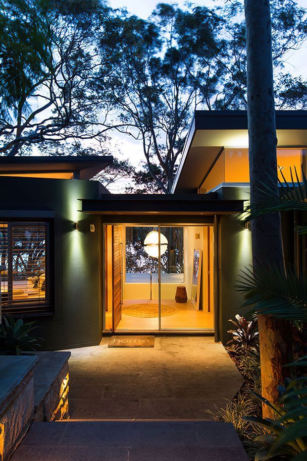 Dusk exterior view of a modern single-storey home entrance set among mature trees and dense planting. The dark rendered facade has broad flat roof planes, integrated exterior lighting, large glazed entry doors, and a warm, welcoming interior visible through the doorway. Concrete or stone steps and paving lead up to the entrance.