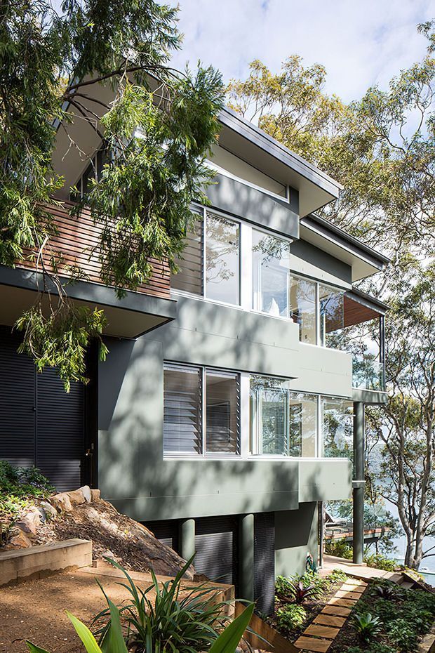 A multi-level contemporary house set into a steep, tree-covered site, viewed from outside. The home features muted grey-green rendered wall panels, extensive glazing, dark louvred openings, horizontal timber cladding accents, projecting roof planes, and elevated structural supports. A narrow stepped path and landscaped garden descend around the house, with glimpses of water and surrounding trees through the glazing.