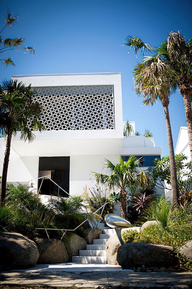 Exterior view of a white, rectilinear modern home set within a lush tropical landscape. The facade features a prominent perforated geometric breeze-block screen, flat rooflines, white rendered walls, dark recessed entry, and a stepped concrete path bordered by rocks, palms, and ornamental planting.