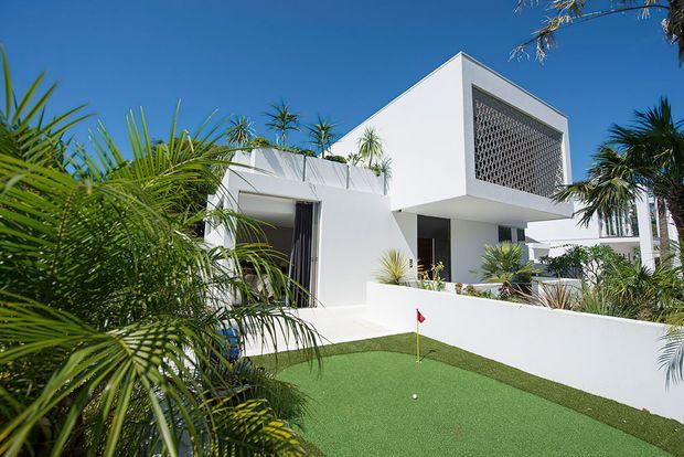Exterior view of a crisp white, flat-roofed modern residence surrounded by tropical landscaping. The home features strong geometric volumes, deep overhangs, large openings, and a decorative breeze-block screen on the upper facade. A small private putting green occupies the foreground.