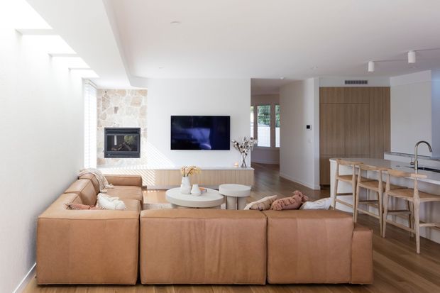 Bright open-plan living room with a large tan sectional sofa facing a wall-mounted television and stone-clad fireplace. The space opens to a contemporary kitchen with a light island benchtop, timber cabinetry, and stools. White walls, pale timber flooring, skylights, and abundant natural light create an airy, refined interior.