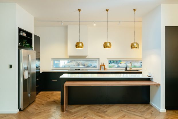 Contemporary kitchen with a large black island clad in dark cabinetry and topped with a light benchtop, paired with a warm timber dining or breakfast counter. The space features herringbone timber flooring, white walls, integrated horizontal windows, stainless steel refrigeration, black shelving, and three brass pendant lights.