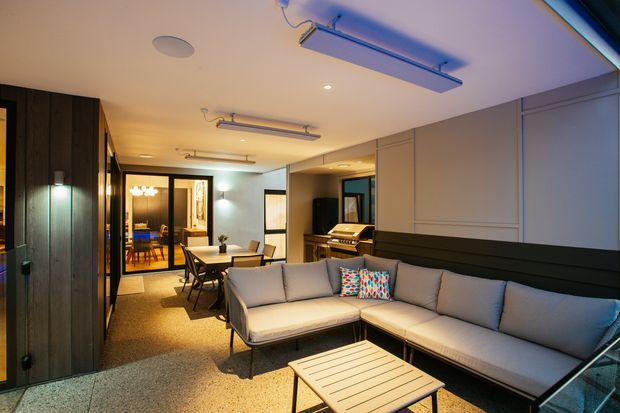 A contemporary open-plan living and dining area with a large grey sectional sofa, light wood coffee table, dining table and chairs, and a piano set into a recessed alcove. Dark timber wall panelling and cabinetry contrast with pale feature wall panels, while linear ceiling-mounted lights and a polished concrete or terrazzo-style floor create a modern, entertainment-focused interior.