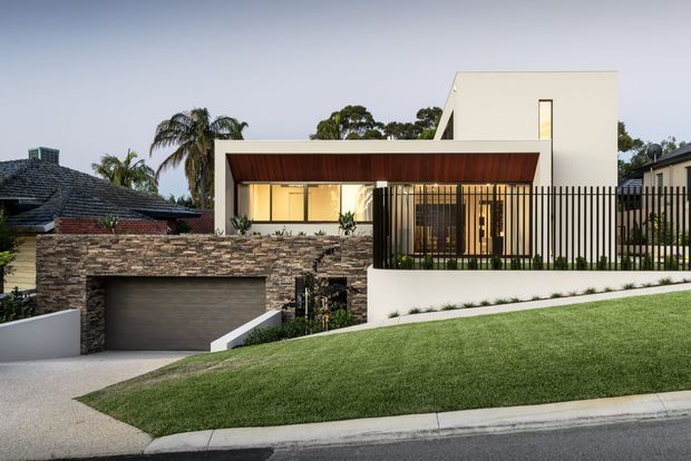 Contemporary residential house exterior at dusk, viewed from the street on a gently sloping site. The facade combines smooth white rendered volumes, a warm timber-lined recessed entry or living area, extensive glazing, dark vertical metal fencing, and a layered natural-stone retaining wall. A wide integrated garage sits below the house, with manicured lawn and neighboring homes visible around it.