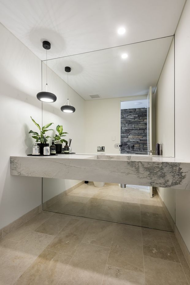 Contemporary bathroom vanity with a full-width frameless mirror, floating pale stone benchtop, wall-mounted basin tapware, pendant lighting, and large beige stone-look floor tiles. A stacked-stone feature wall is visible through the doorway, with potted greenery and bathroom accessories on the vanity.