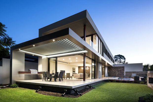 A contemporary two-storey residence photographed from the rear garden, featuring strong rectilinear forms, extensive floor-to-ceiling glazing, a covered outdoor entertaining terrace, and a projecting louvred pergola. The neutral rendered facade is paired with dark cladding, slim dark window frames, stone feature walls, and a landscaped lawn.