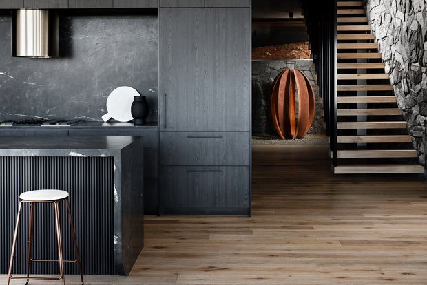 A dramatic contemporary kitchen and stair hall featuring dark grey textured cabinetry, a dark stone-look splashback, a ribbed island panel, warm timber flooring, and a sculptural timber-and-black stair. A rough stacked-stone wall and black metal detailing add a refined industrial character, while a single bar stool and minimal decorative objects complete the scene.