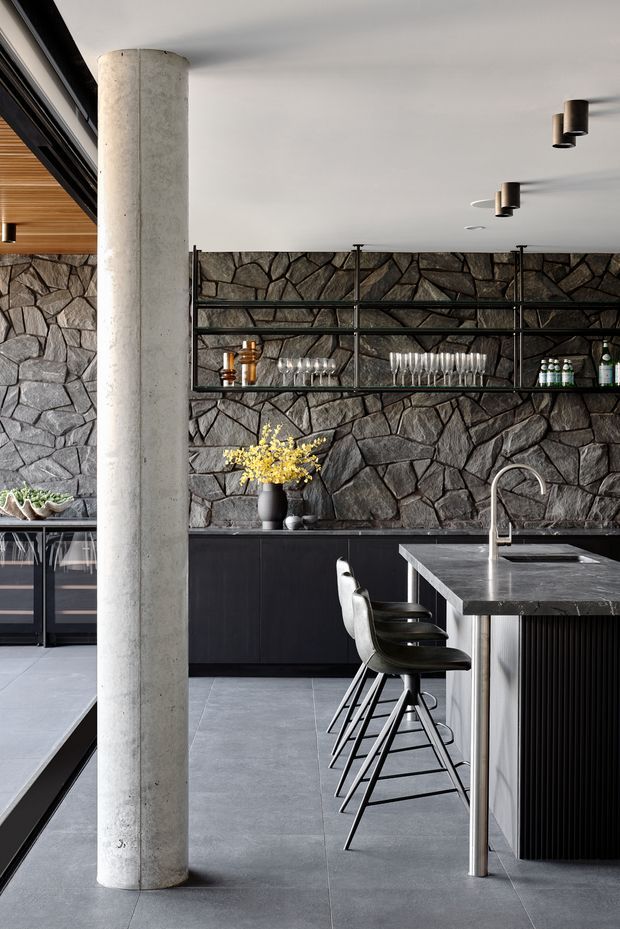 A sophisticated contemporary kitchen with a dark, textural irregular stone feature wall, slim black open shelving, matte black cabinetry, and a large dark stone island with integrated sink and gooseneck tap. Black bar stools line the island. A cylindrical raw concrete column stands prominently in the foreground, while timber ceiling lining, black-framed glazing, concrete-look flooring, and minimalist cylindrical pendant lights reinforce the refined industrial character.