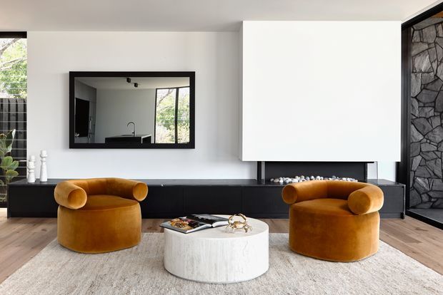 A contemporary living room with two sculptural mustard velvet lounge chairs facing a low round white coffee table and a long black built-in media console. A large wall-mounted white projection screen or artwork dominates the wall, with a black-framed mirror reflecting the adjacent space. Light timber flooring, a pale textured rug, floor-to-ceiling glazing, and a dark irregular-stone feature wall create a refined, high-contrast interior.