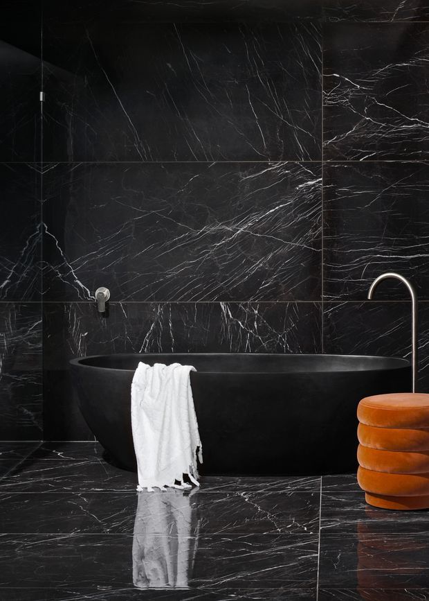 A dramatic, luxurious bathroom dominated by black marble-look wall and floor surfaces with fine white veining. A matte black freestanding oval bathtub sits against the tiled wall, accompanied by a curved dark metal floor-mounted faucet. A white towel hangs over the tub, and a sculptural burnt-orange upholstered stool adds a warm accent. The composition is high-contrast and refined, with glossy floor reflections.