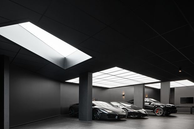 A dark, highly stylised indoor car showroom or display garage with several luxury sports cars arranged beneath large illuminated ceiling panels and skylight-like light wells. The space has charcoal walls and ceiling, exposed square structural columns, a smooth grey floor, and sparse display elements including a wall-mounted screen. The composition emphasises dramatic architectural lighting, clean lines, and a premium automotive setting.