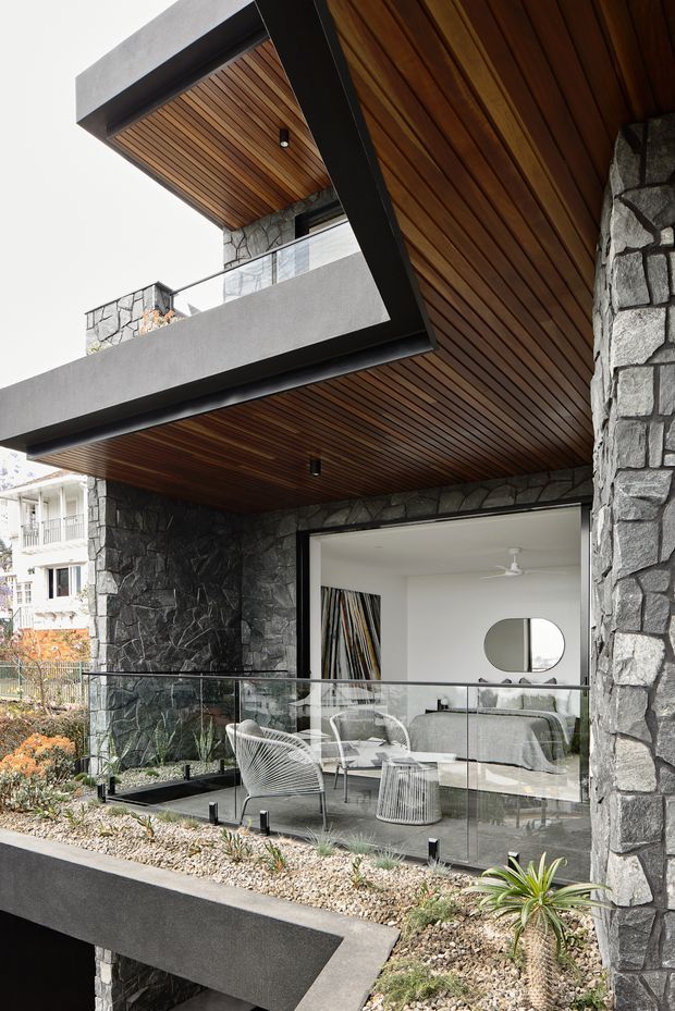Exterior architectural view of a contemporary multi-level residence with dark rough stone walls, charcoal concrete or rendered structural beams, extensive timber-lined soffits, and a glazed balcony terrace. A modern living room is visible through the large opening, with pale furniture and a ceiling fan. The image emphasizes the building facade, projecting roof forms, terrace edge, and layered landscaping with gravel and succulents.